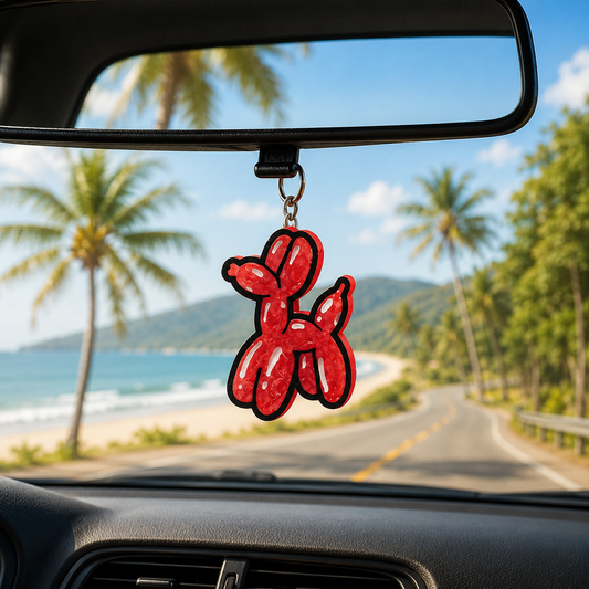 Red balloon dog car air freshener hanging from a car's rearview mirror with a scenic road and beach in the background.