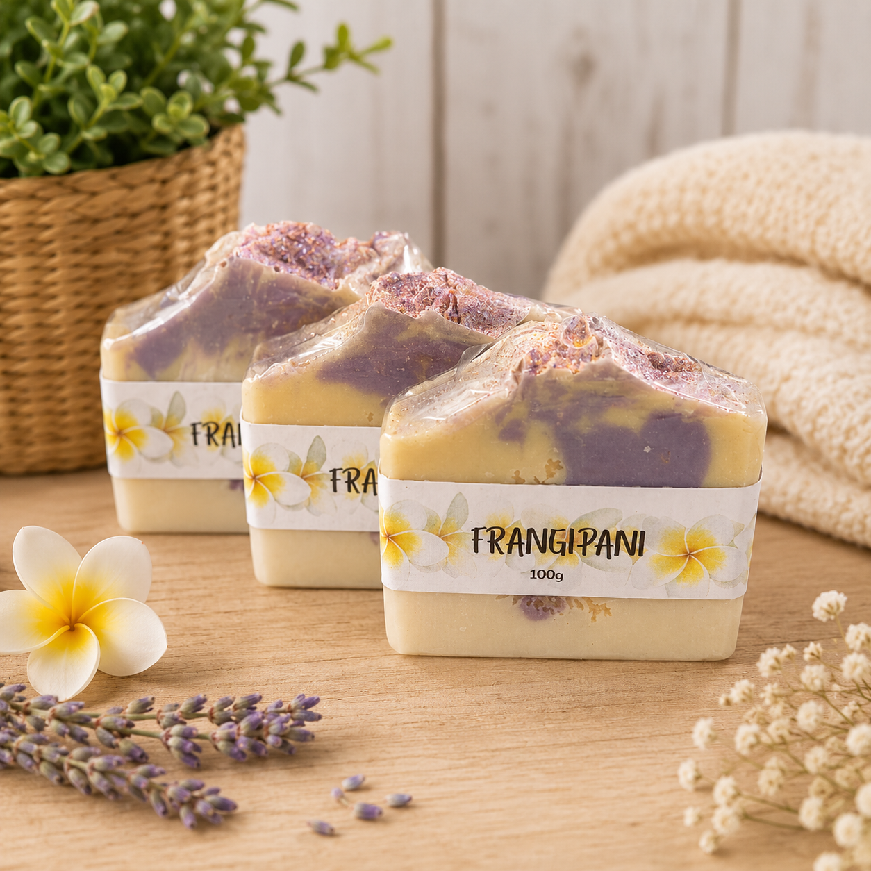 Three bars of soap with 'Frangipani' label on a wooden surface, surrounded by flowers and a plant.