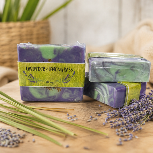 Two bars of lavender lemongrass soap with a label, surrounded by lavender flowers and green leaves on a wooden surface.