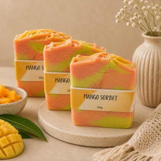 Three bars of mango sorbet soap on a beige surface with a bowl of mangoes and a vase in the background.